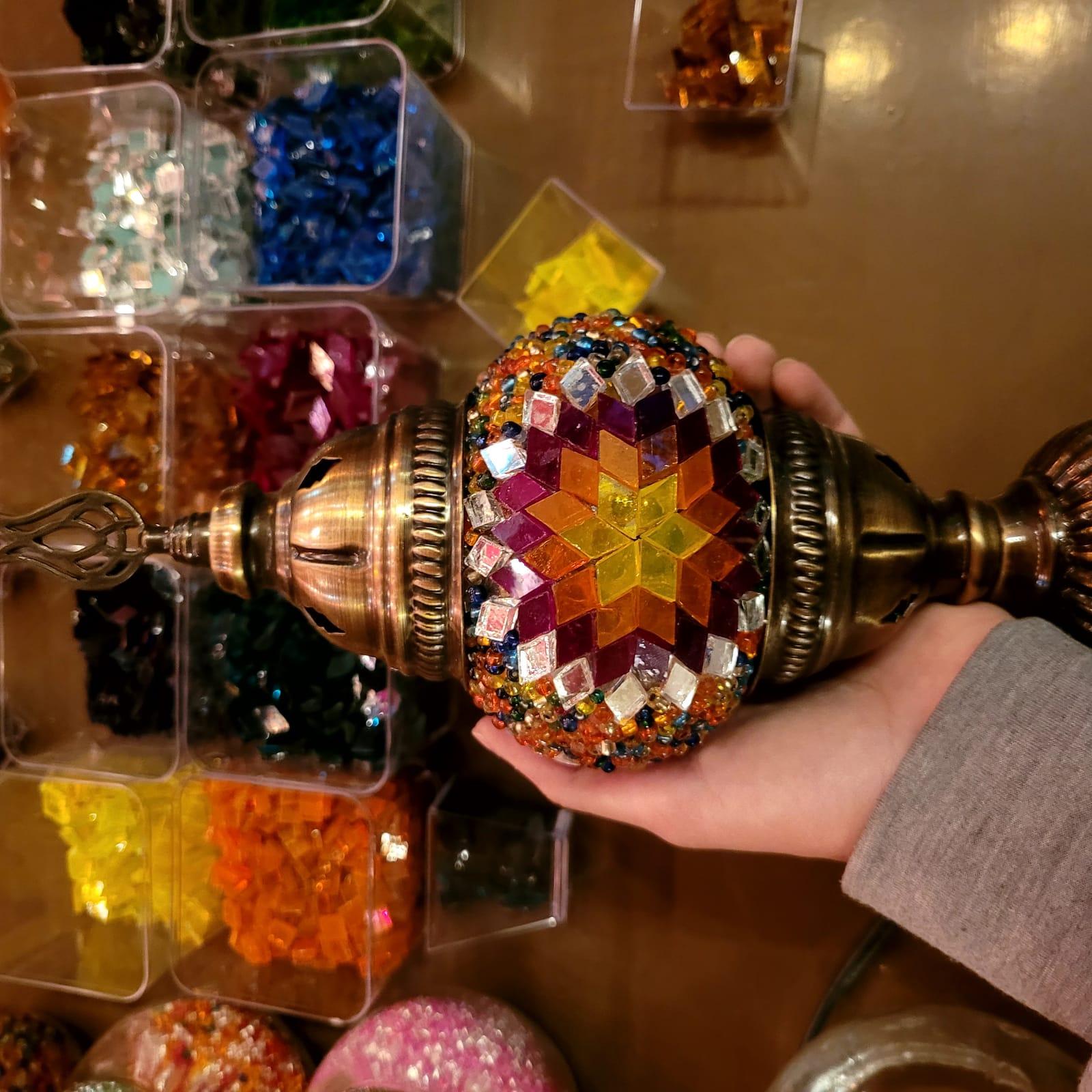 Hands placing mosaic tiles onto a lamp globe, surrounded by glass-tile sorting trays